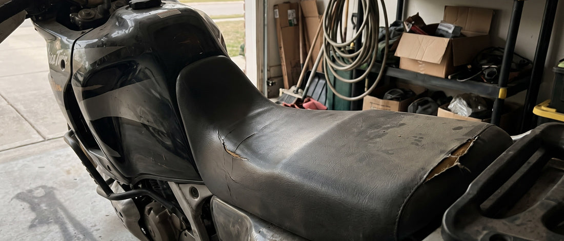 Motorbike seat recover decision: worn Honda Africa Twin seat with cracked factory vinyl at a shaded garage