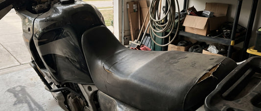 Motorbike seat recover decision: worn Honda Africa Twin seat with cracked factory vinyl at a shaded garage